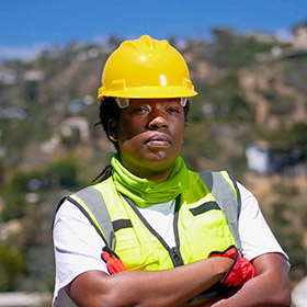 worker-1jpeg - Speedy Gold - Speedy Gold Construction worker wearing a yellow hard hat and high-visibility vest with arms crossed at a construction site.