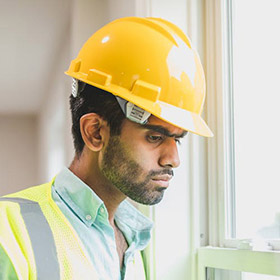 worker-3jpeg - Speedy Gold - Speedy Gold Construction worker wearing a yellow hard hat and reflective vest, looking out a window.