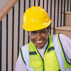 worker-4jpeg - Speedy Gold - Speedy Gold Smiling construction worker in a yellow hard hat and neon safety vest indoors near a staircase.