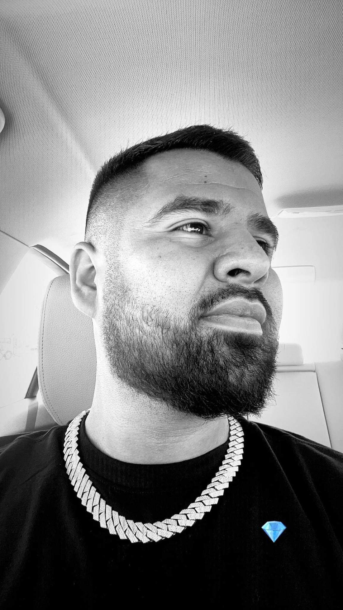 Close-up of a bearded man wearing a chunky silver chain, sitting in a car and looking to the side.