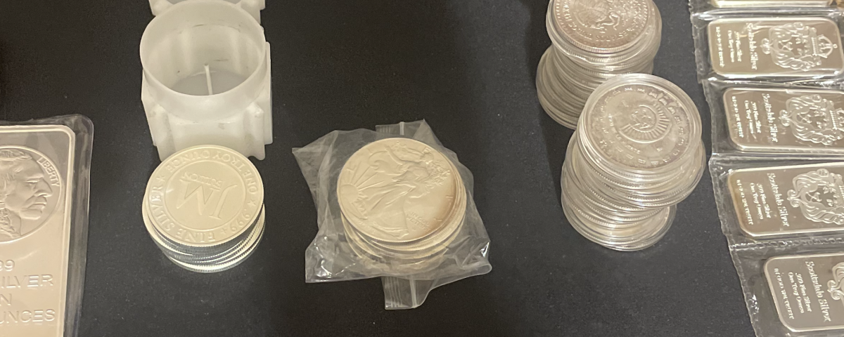 Assorted silver coins and bullion bars on a dark surface, including a large bar on the left and stacked coins across the center and right.