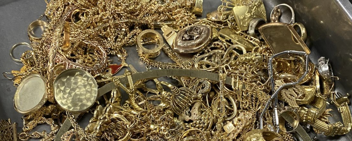 A tangled heap of gold jewelry—chains, bracelets, rings, and watches in a metal container.